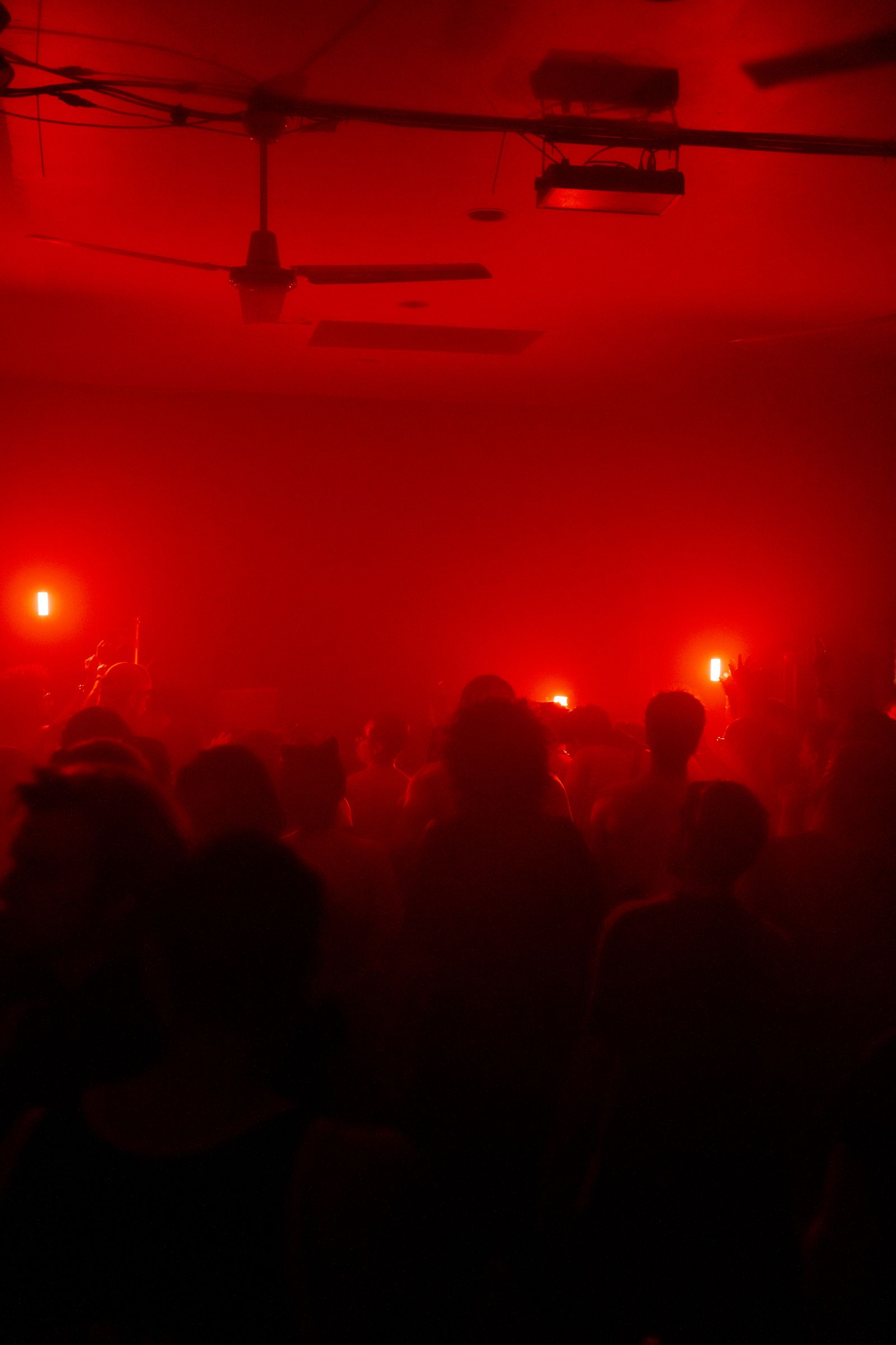 A packed dancefloor in silhouette under red light, haze and ceiling fans