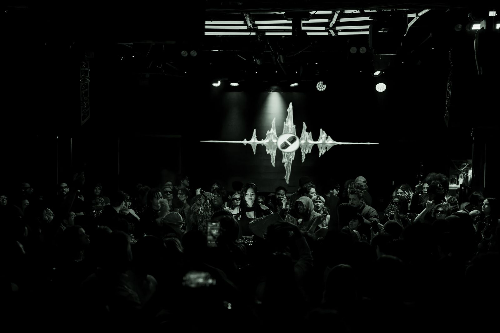 Wide shot of the Brooklyn Monarch floor, crowd packed under a single spotlight as Kronos' jagged waveform sigil glows on the back wall.