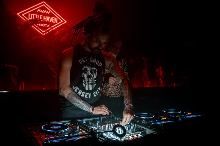 DJ working the CDJs under the Little Haven neon, locs pulled back, headphones around the neck.